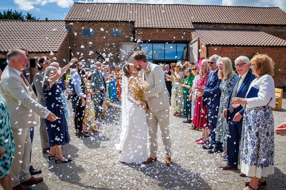 Bride and groom kissing during confetti shower at outdoor wedding ceremony with guests lining the path.