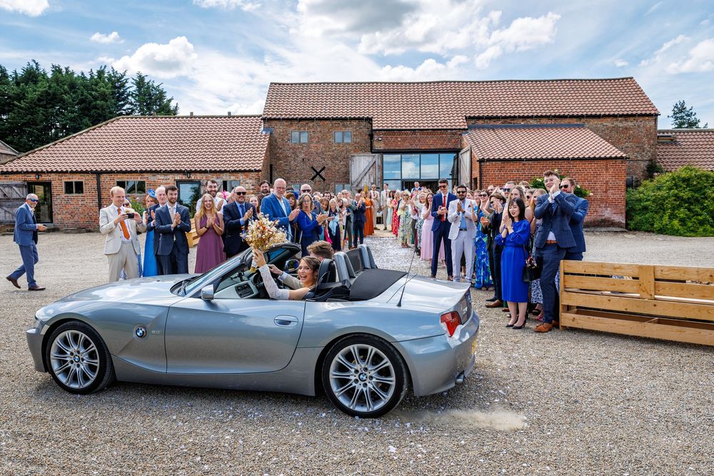Newlyweds depart in silver BMW Z4 convertible as wedding guests celebrate outside a rustic barn venue.