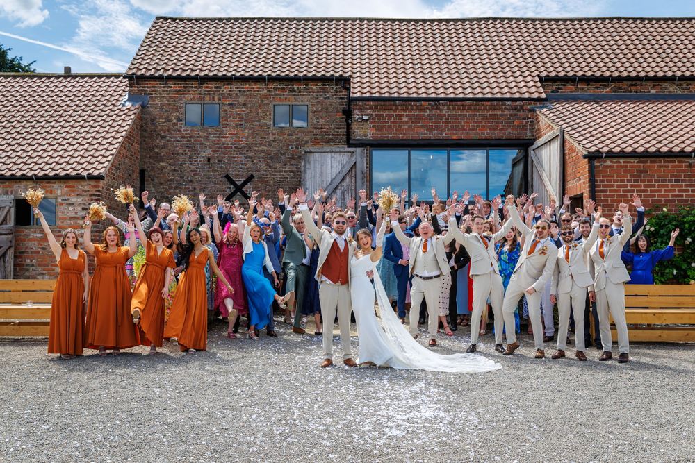A joyful wedding party celebrates outside a rustic barn venue, with the bride and groom surrounded by cheering guests.
