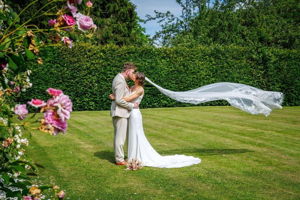 Bride and groom share a romantic kiss in a lush garden, with the wedding veil flowing gracefully in the breeze.
