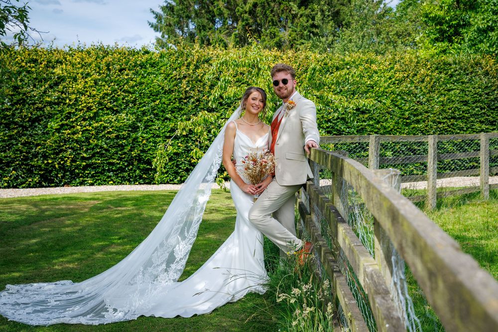 Bride and groom pose by wooden fence in lush garden, bride wearing flowing white gown with long veil.