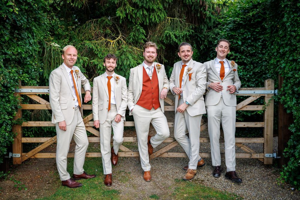 Five groomsmen in cream suits with rust orange ties pose by a wooden gate surrounded by lush green ivy.