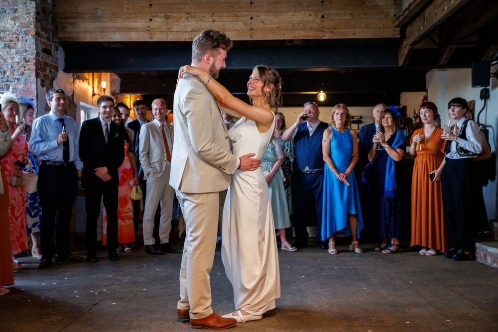 Bride and groom share first dance at rustic wedding reception with guests watching in background.