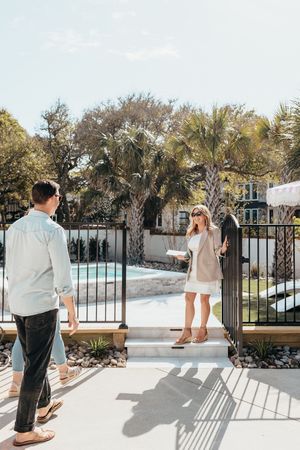 A woman opens a pool gate for a man at a sunny outdoor pool area surrounded by palm trees.