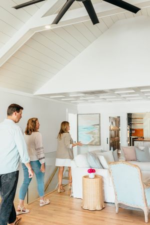 Three people touring a bright coastal living room with vaulted ceiling, ceiling fan, and light blue decor.