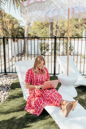 Woman in red floral dress working on pink laptop outdoors on white lounger under striped fringe umbrella.