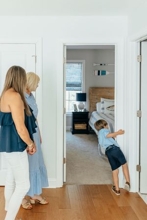 Two women and a young boy exploring a coastal-decorated bedroom doorway in a bright, modern home.