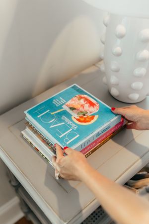 Woman with red nails arranging a stack of books including Beach Cocktails on a white side table.