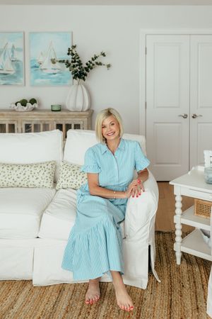 Smiling blonde woman in a light blue striped dress sitting on a white slipcovered sofa in a coastal-themed living room.