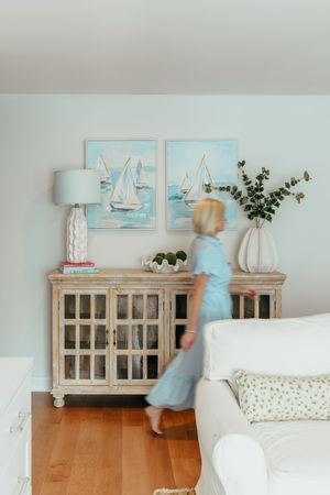 Coastal living room with sailboat paintings, rustic cabinet, white lamp, and woman in blue dress walking past.