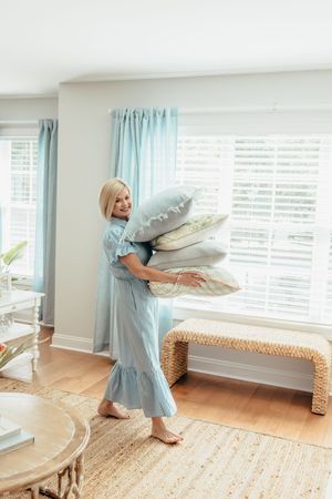 Woman in blue dress carrying stack of decorative pillows in bright, coastal-style living room with woven bench.