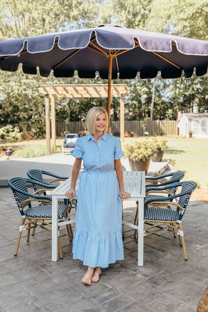 Woman in light blue midi dress standing by white patio table under navy umbrella in elegant outdoor setting.