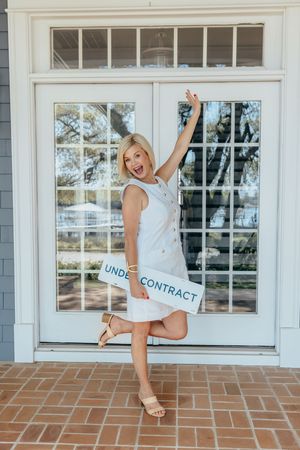 Excited blonde woman in white dress holding an Under Contract real estate sign on a brick porch.