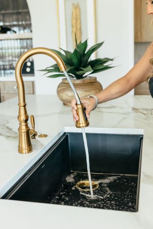 A hand uses a gold pull-down kitchen faucet over a dark farmhouse sink on a white marble countertop.