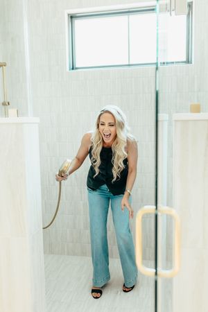 Laughing blonde woman in black top and jeans holding a shower head inside a modern tiled walk-in shower.