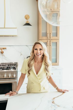 Smiling blonde woman in yellow blazer dress leaning on marble kitchen island in modern white kitchen.