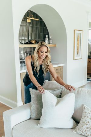 Blonde woman arranging decorative pillows on a gray sofa in a modern living room with an arched built-in bar.