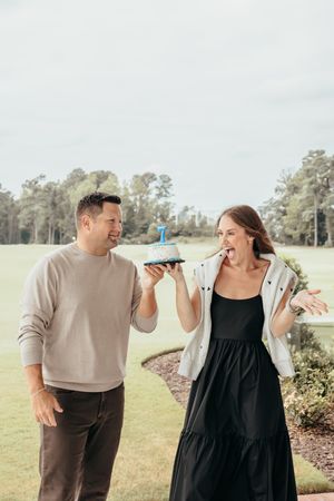 A couple smiling outdoors holding a small birthday cake with a candle on a golf course.