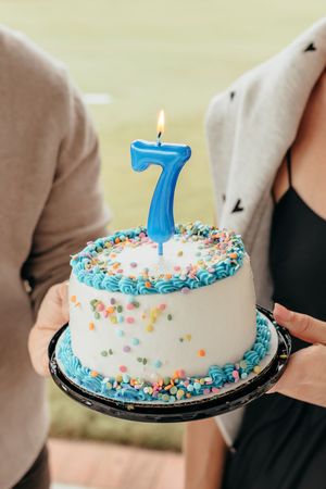 A white birthday cake with blue frosting, colorful sprinkles, and a lit blue number 7 candle held by two people.