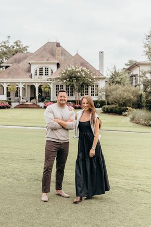 A couple stands smiling on a green lawn in front of a large elegant white home with lush landscaping.