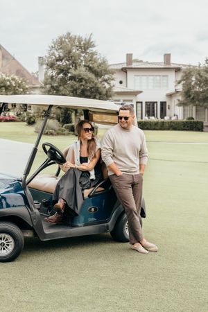 A stylish couple in sunglasses relaxing by a blue golf cart on a lush green golf course near an elegant clubhouse.