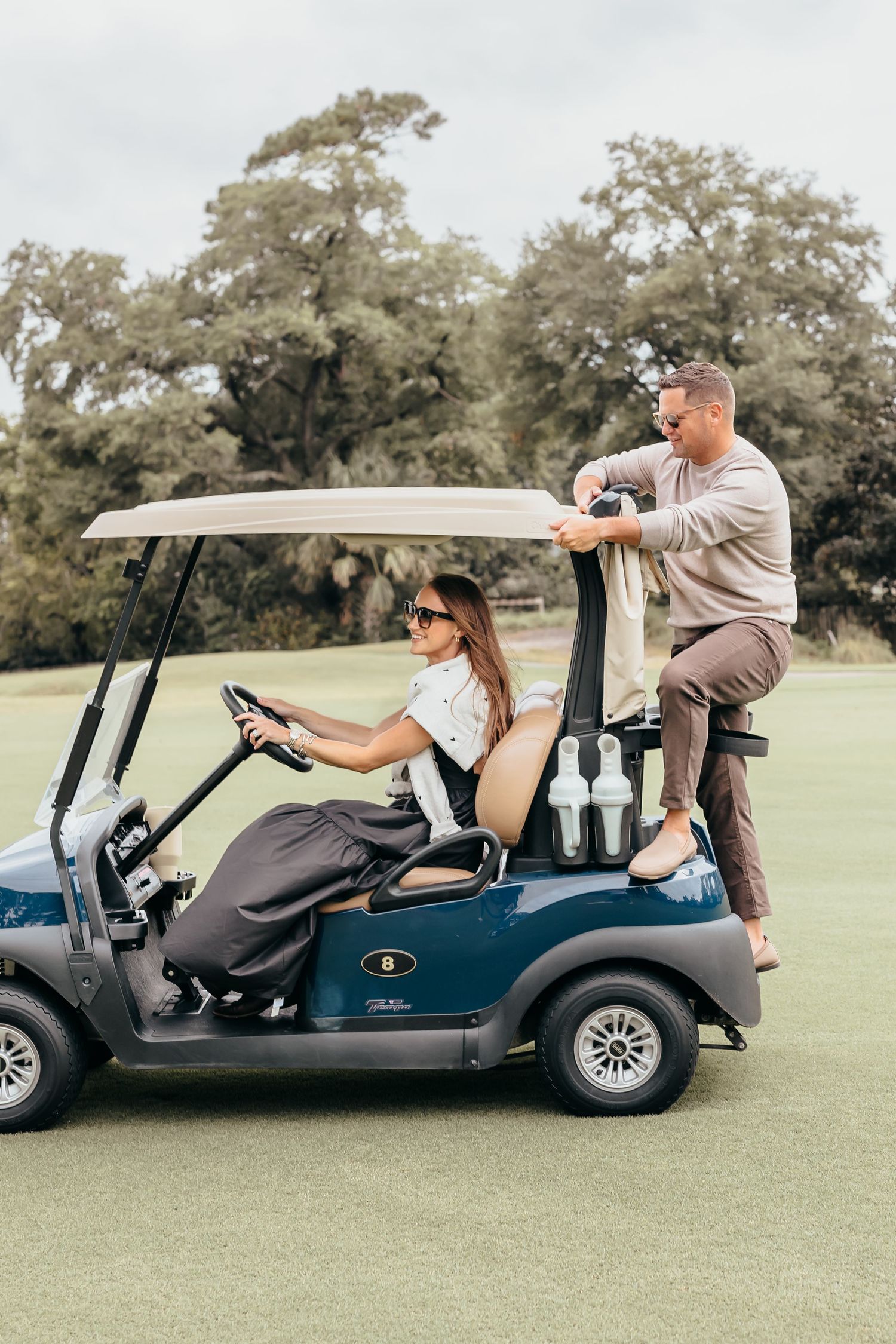 A woman in sunglasses drives a blue golf cart on a green course while a man leans on the back chatting.