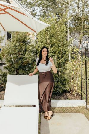 Woman in white top and brown maxi skirt poses by pool lounge chair under patio umbrella in sunny backyard.