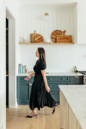 Woman in black pleated dress walking through modern kitchen with teal cabinets and open wooden shelving.