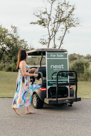 Woman in floral dress beside a Nest Realty branded golf cart with a For Sale sign outdoors.