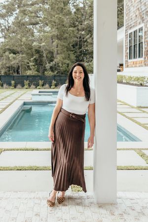 Woman in white top and brown pleated maxi skirt posing by a modern pool patio with lush greenery.