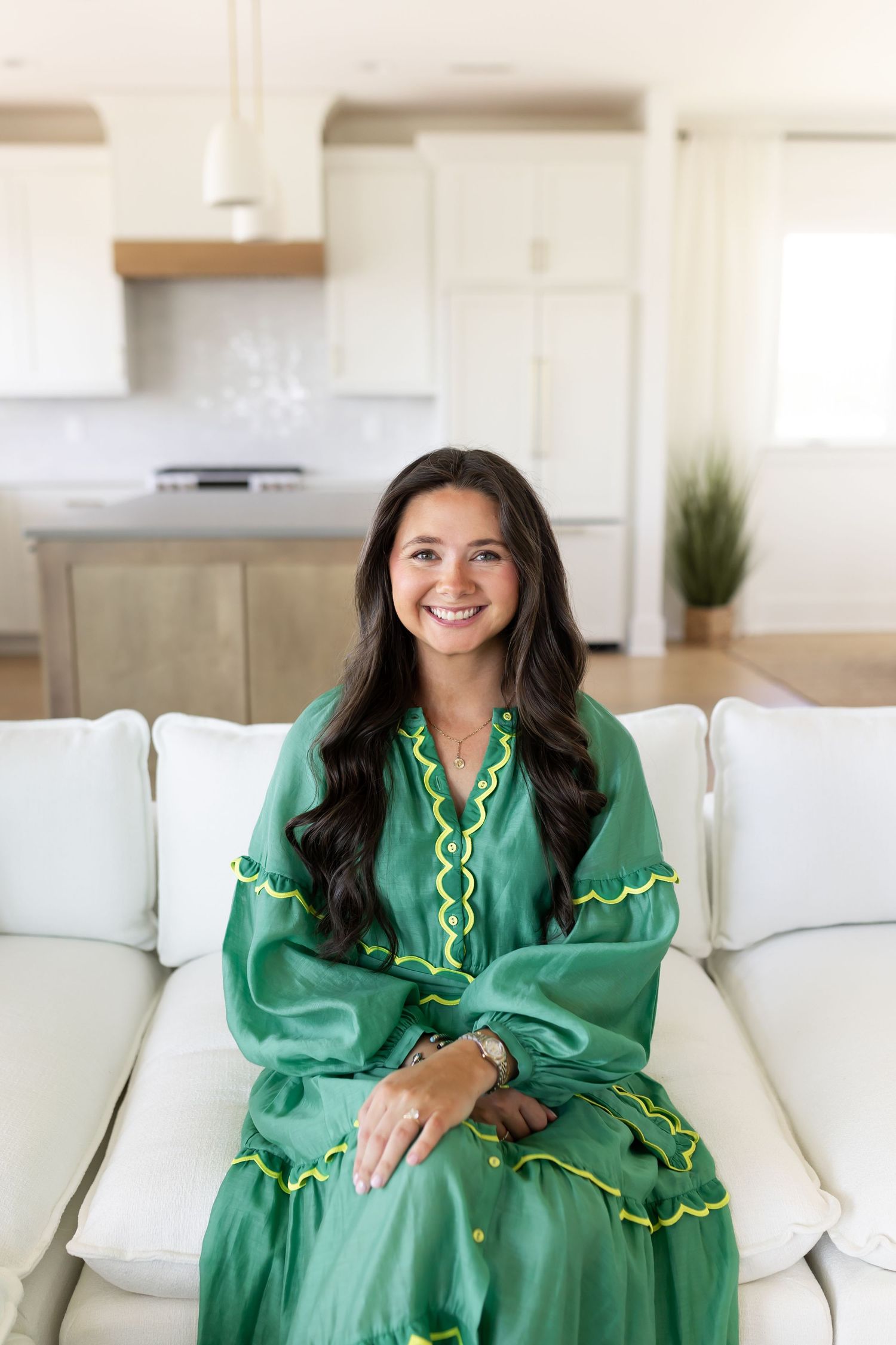 Woman in green embroidered dress sitting on white sofa, smiling in modern kitchen background.