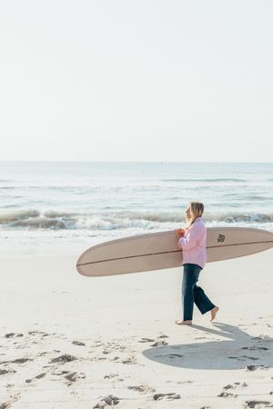 A woman in a pink sweater and jeans carries a longboard surfboard along a sandy beach near the ocean.