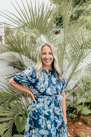 Smiling blonde woman in a blue floral dress posing in front of lush tropical palm leaves outdoors.