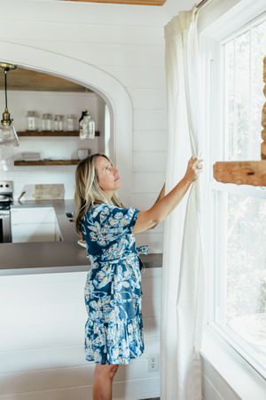 Woman in blue floral dress adjusting white curtains by a bright window in a modern farmhouse kitchen.