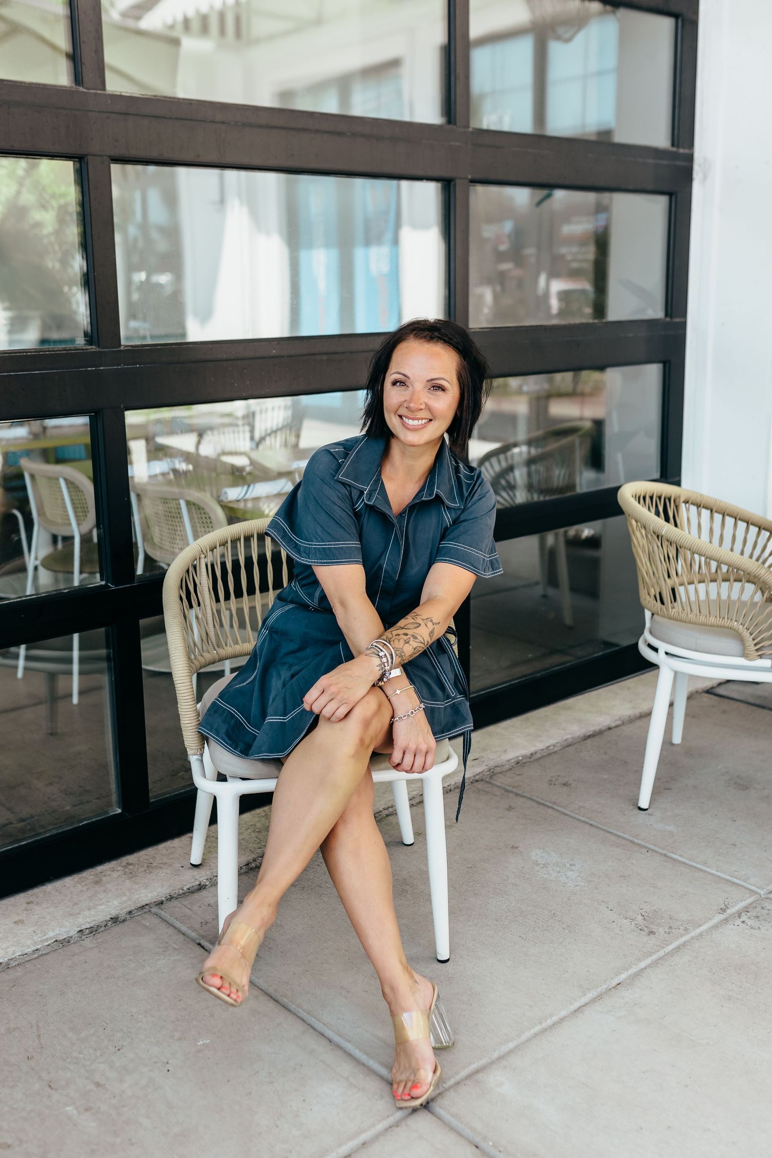 Woman in denim dress sitting on white chair, smiling outdoors near large windows.