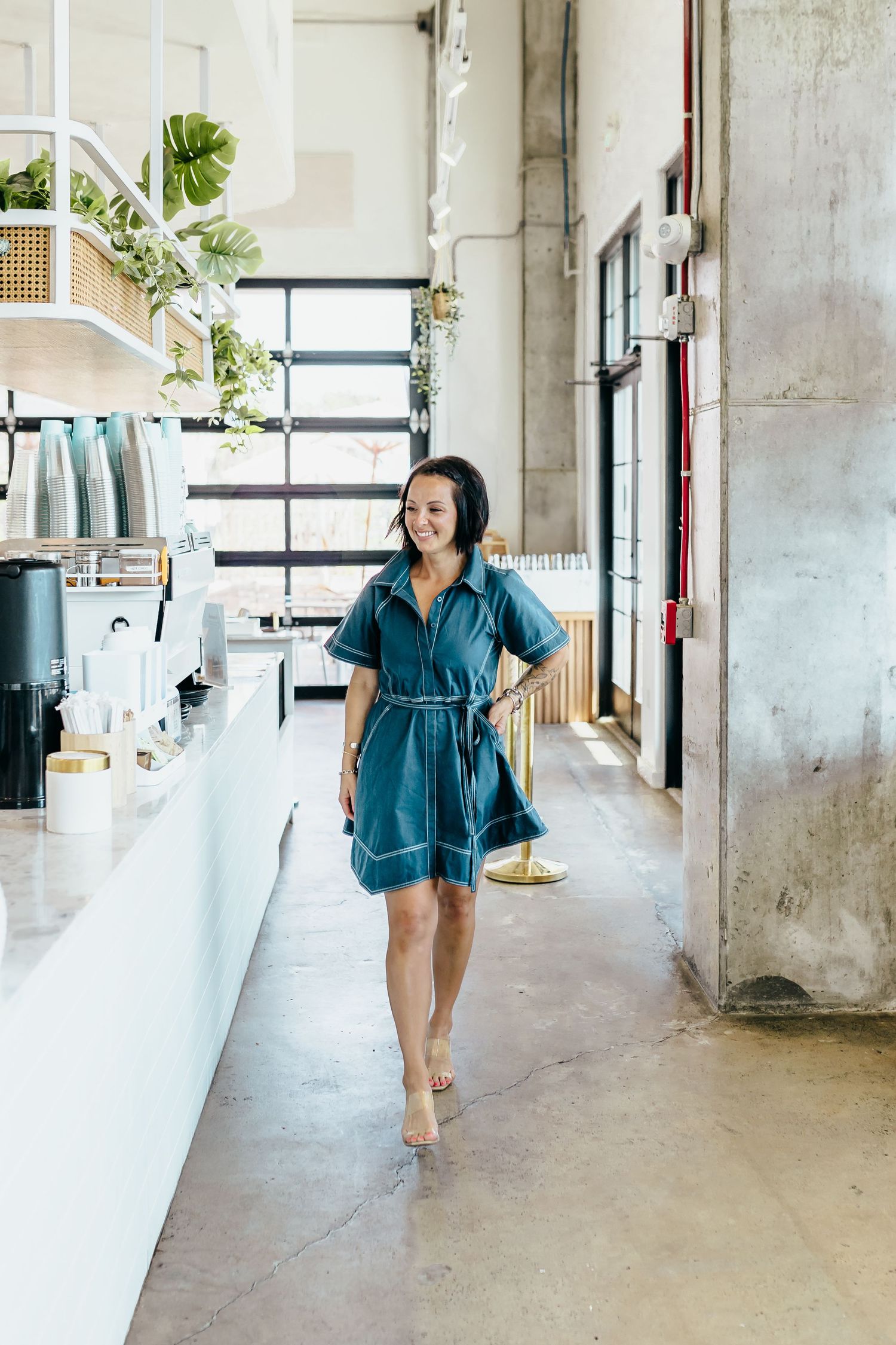 Woman in teal shirt dress walking through a modern cafe with concrete floors and lush green plants.