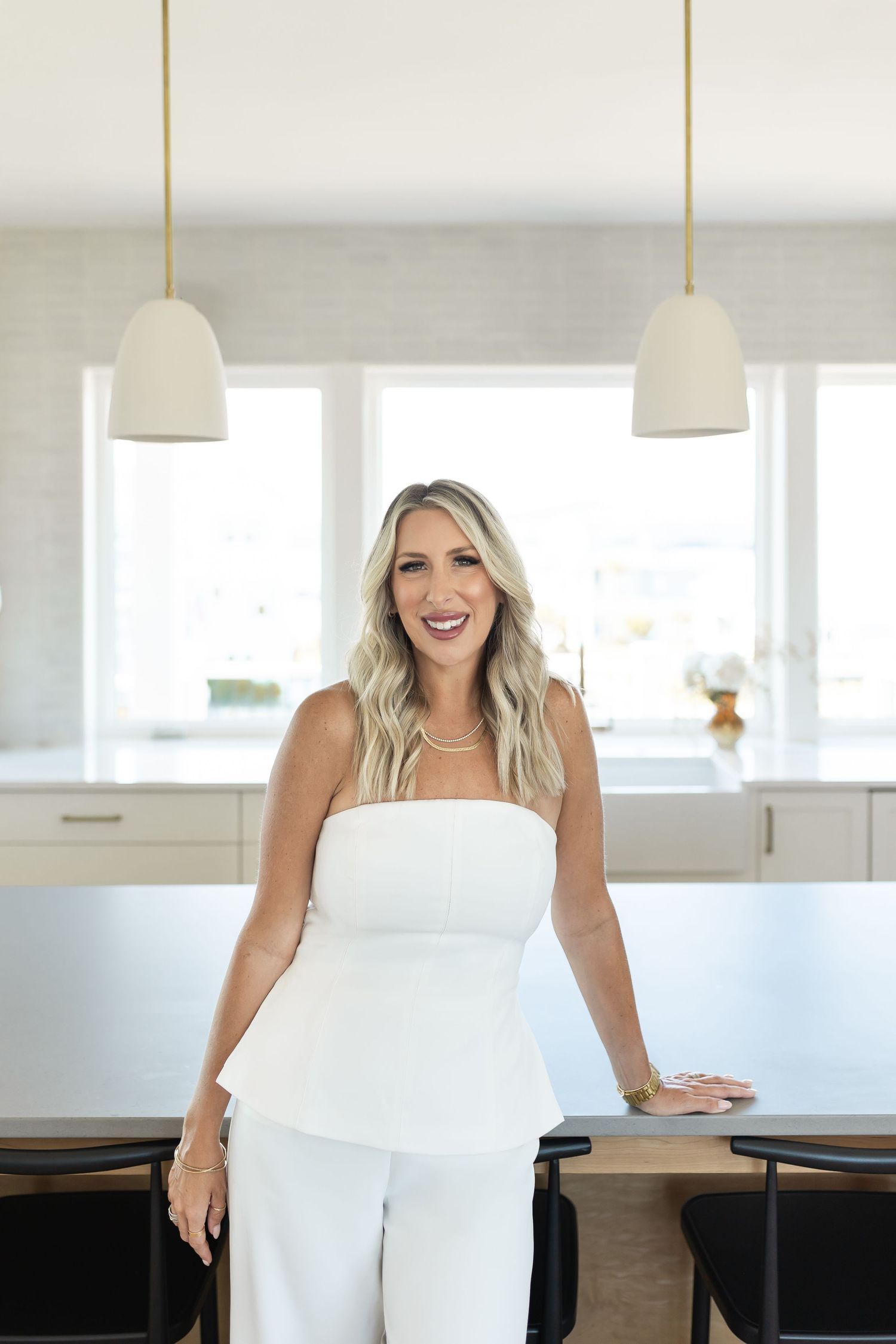 Smiling blonde woman in white strapless peplum outfit standing in a modern white kitchen with pendant lights.