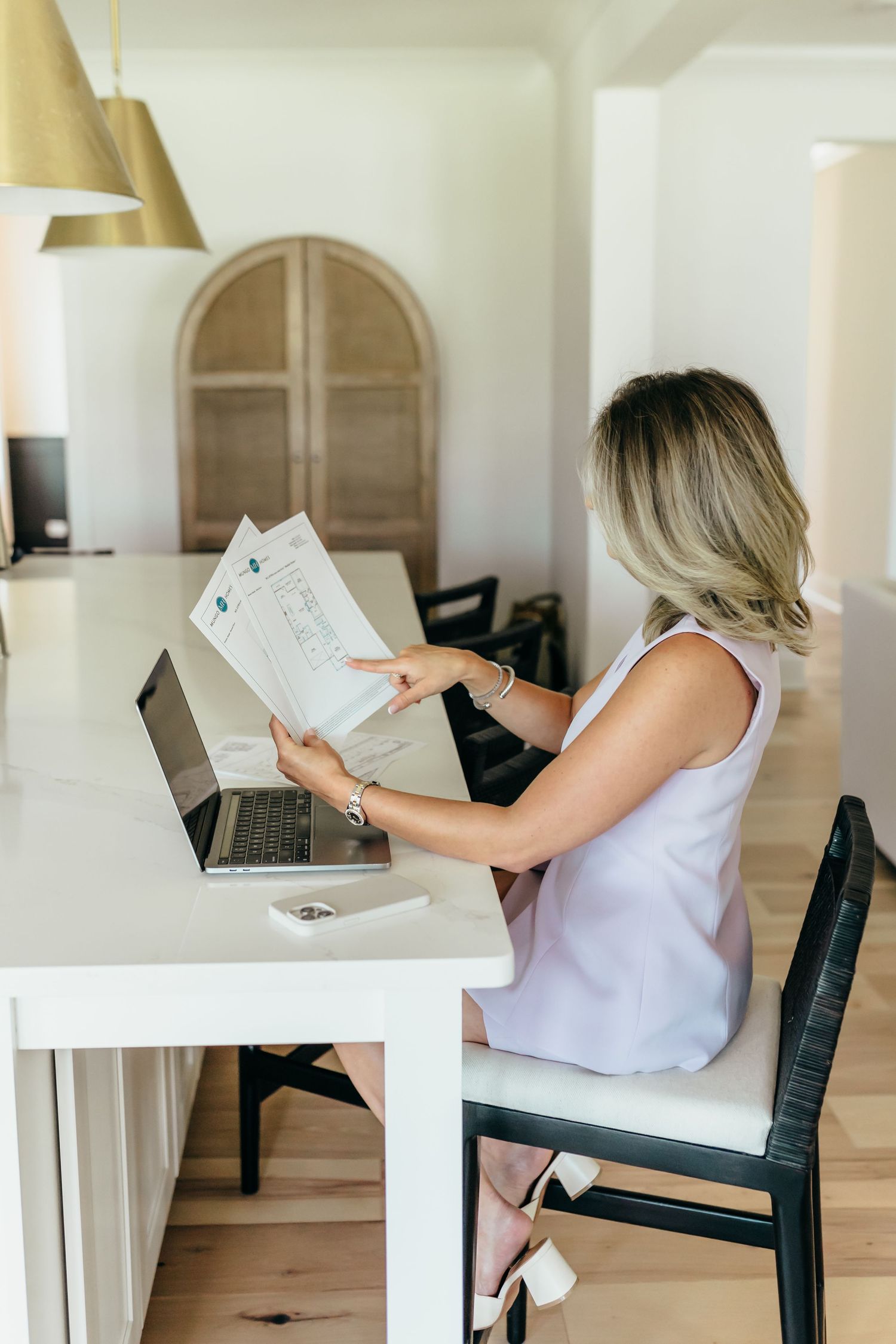 Woman reviewing documents at white desk with laptop in bright home office setting.