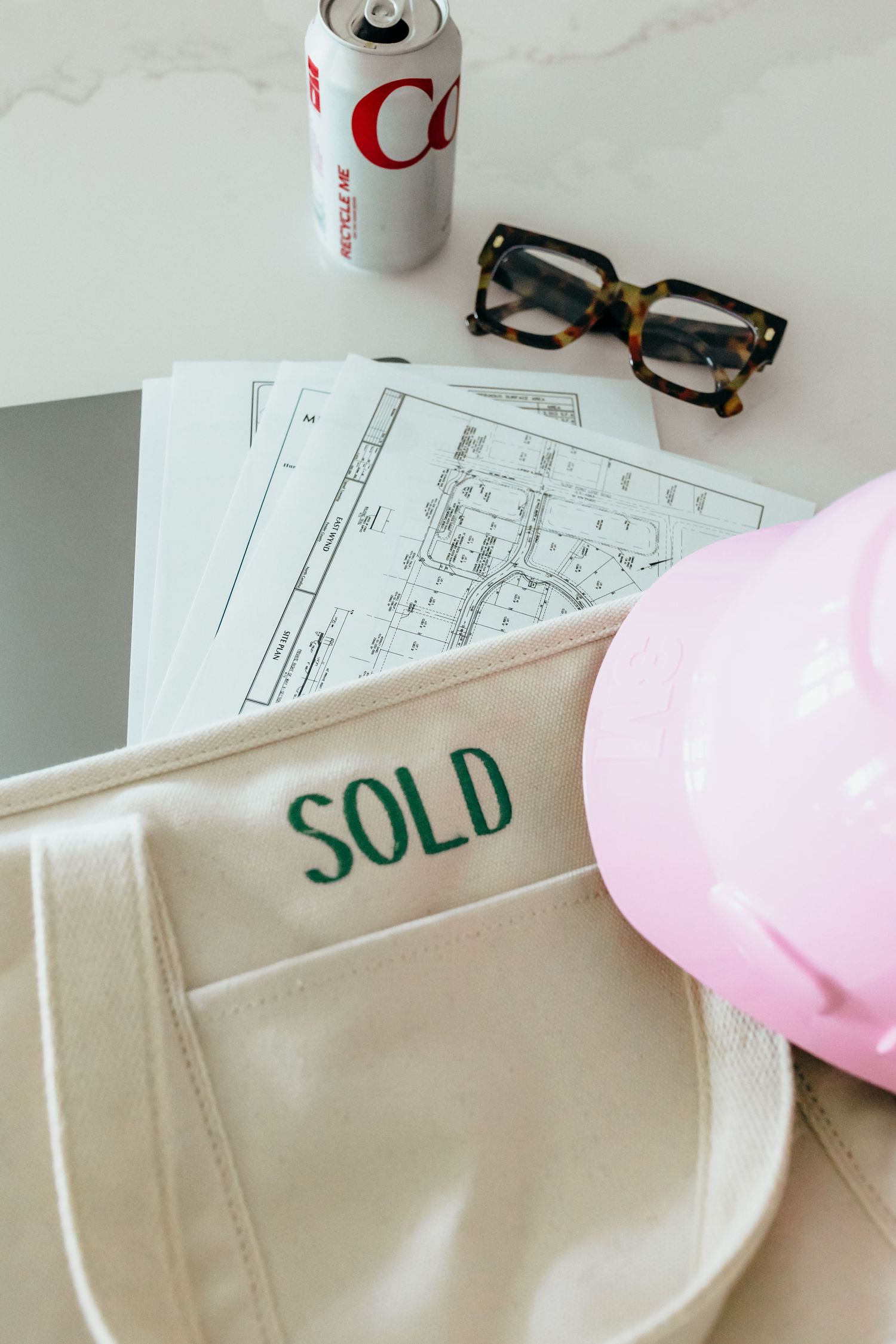 A cream tote bag embroidered with SOLD sits beside blueprints, tortoise glasses, a soda can, and a pink hard hat.