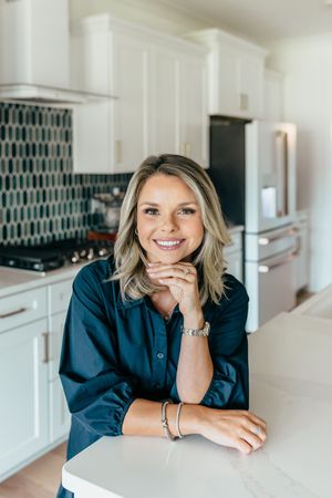 Smiling blonde woman leaning on white kitchen counter with navy blouse, bracelet, and teal geometric backsplash.