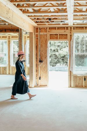Woman in black dress and pink hard hat walking through a wood-framed house under construction.