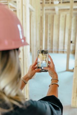 Woman in pink hard hat photographs construction site interior framing with smartphone.