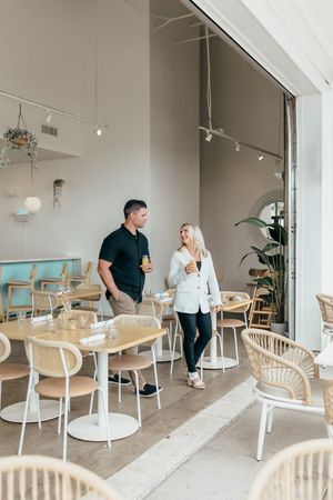 A couple walks through a modern cafe with rattan chairs, holding drinks and smiling at each other in a bright, airy space.