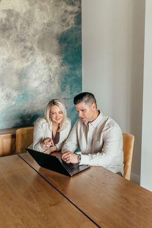 A man and woman sit at a wooden table, smiling while collaborating on a laptop in a modern room.