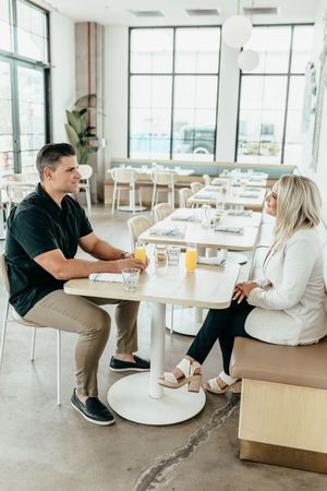 A man and woman sit across from each other at a light wood table in a bright, modern restaurant with orange juice glasses.