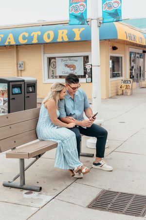 Couple sitting on a bench outside a beachside factory store, looking at a smartphone together on a sunny day.