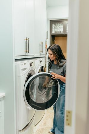Woman smiling while loading laundry into a front-load washing machine in a modern white laundry room.
