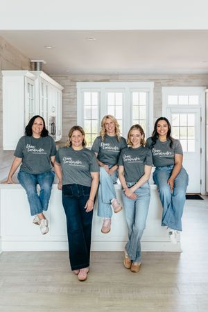 Five women in matching gray branded t-shirts and jeans smiling together in a bright modern kitchen.