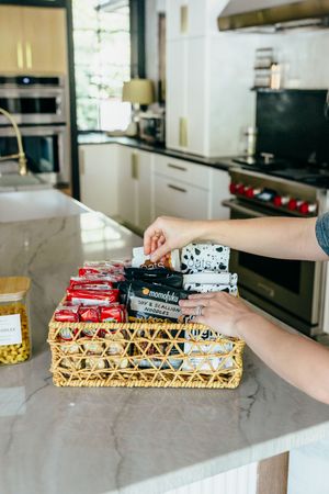 Hands arranging snacks in a wicker basket on a marble kitchen counter with modern appliances in background.
