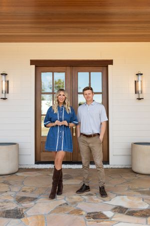 A man and woman stand smiling in front of elegant double wood doors on a stone patio of a modern farmhouse.